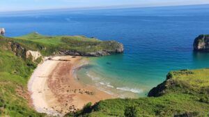 alt="Vista de la Playa de Ballota en Llanes, Asturias, con arena dorada y el islote de Castro Ballota en el mar.">